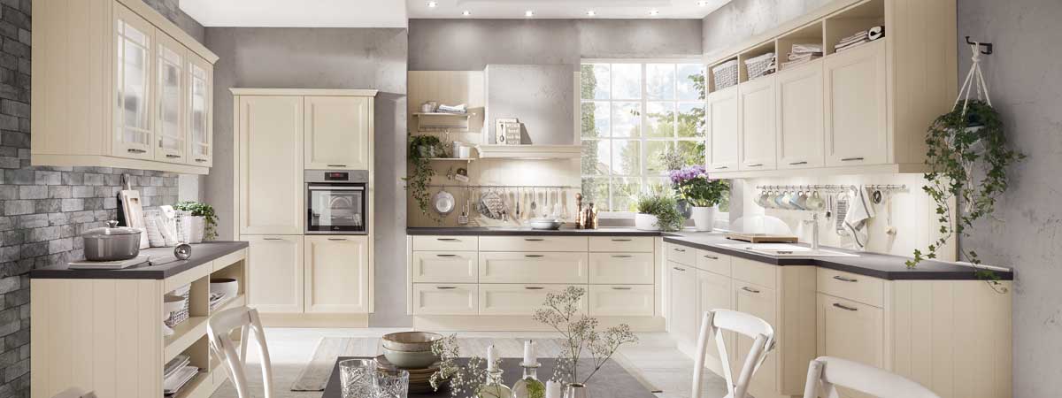 Helle Landhausküche mit cremefarbenen Holzmöbeln, viel Stauraum, großen Fenstern, Pflanzen und gemütlicher Atmosphäre.